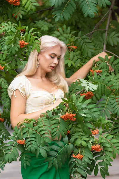 Sarı korseli genç bir kadın, çıplak göbekli ve yeşil bir midi-sarılı etekli, bir Rowan ağacının yanında.