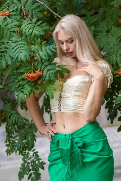 Sarı korseli genç bir kadın, çıplak göbekli ve yeşil bir midi-sarılı etekli, bir Rowan ağacının yanında.