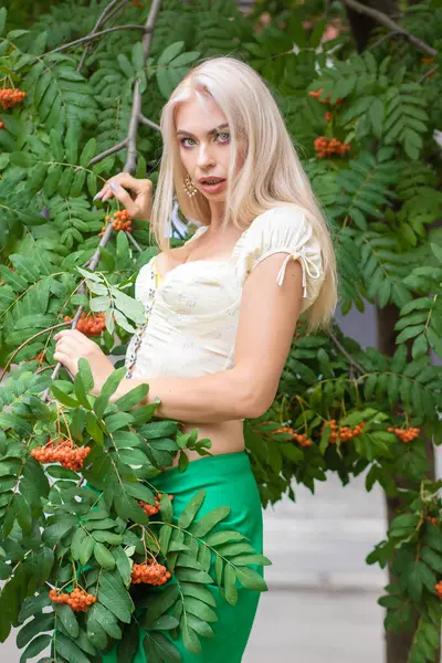 Sarı korseli genç bir kadın, çıplak göbekli ve yeşil bir midi-sarılı etekli, bir Rowan ağacının yanında.