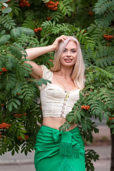 Sarı korseli genç bir kadın, çıplak göbekli ve yeşil bir midi-sarılı etekli, bir Rowan ağacının yanında.