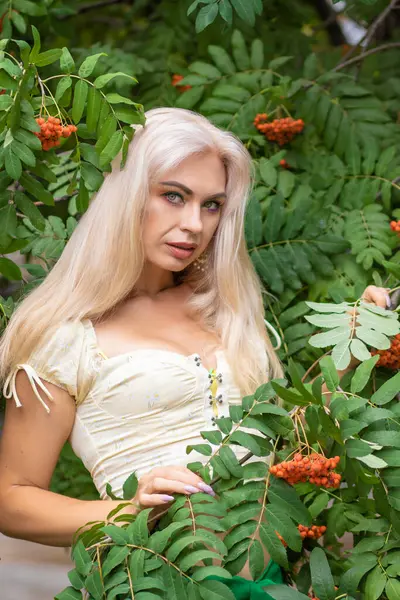 Sarı korseli genç bir kadın, çıplak göbekli ve yeşil bir midi-sarılı etekli, bir Rowan ağacının yanında.