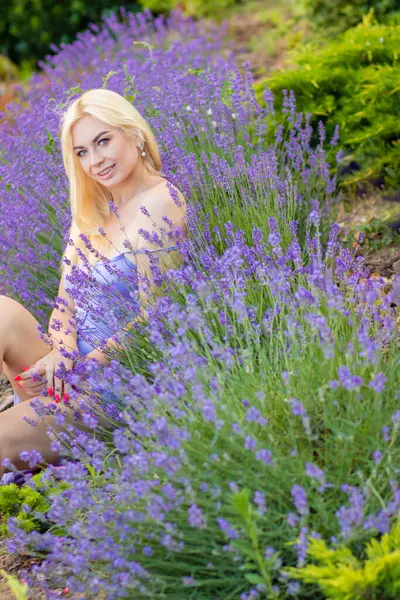 Leylak rengi bir elbisenin içinde uzun saçlı ve doğal makyajlı bir sarışının portresi lavanta çiçeklerinin arasında bir tarlada duruyor. Yaz sabahı