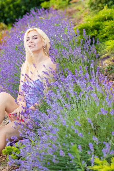 Leylak rengi bir elbisenin içinde uzun saçlı ve doğal makyajlı bir sarışının portresi lavanta çiçeklerinin arasında bir tarlada duruyor. Yaz sabahı