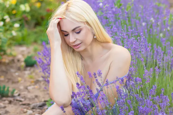 Leylak rengi bir elbisenin içinde uzun saçlı ve doğal makyajlı bir sarışının portresi lavanta çiçeklerinin arasında bir tarlada duruyor. Yaz sabahı
