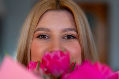 Bir buket pembe lalenin arkasına saklanan genç bir kadının yakın plan portresi.