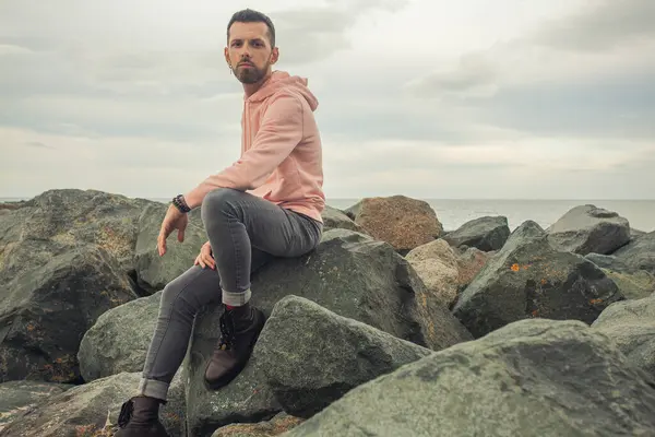 Männermodekonzept Junger Bärtiger Gutaussehender Mann Sitzt Trendiger Kleidung Steinigen Meeresufer — Stockfoto