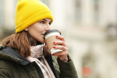 Kahve aşığı konsepti. Avrupa 'nın caddesinde sıcak içecek içen genç bir kadının portresi. Yeşil manken, beyaz ve bej eşarp, sarı bere. Kapatın. Açık hava çekimi