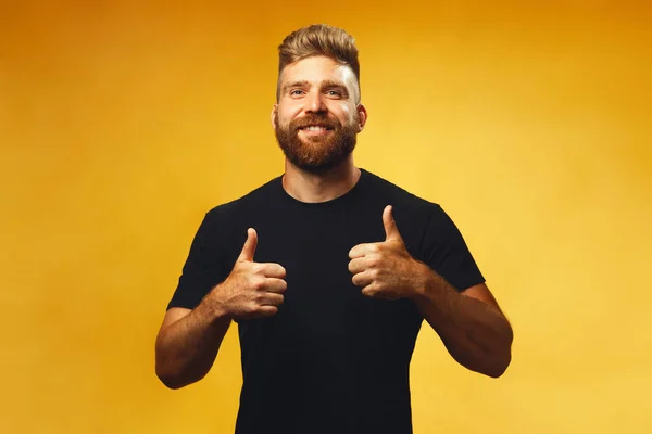 Male beauty concept. Portrait of happy, smiling 35-year-old man with fit body posing over yellow background in black t-shirt, showing thumbs up. Red hair, modern haircut. Copy-space. Studio shot