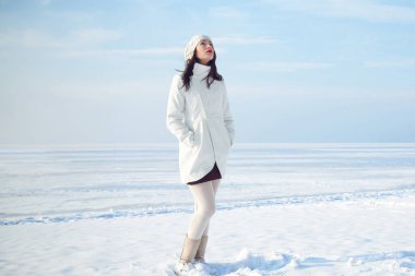 Kış moda konsepti. Kar kıyısında duran beyaz önlüklü ve bereli modaya uygun bir modelin tam boy portresi. Güneşli bir hava. Fransız tarzı. Metin uzayı. Açık hava çekimi.