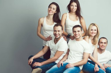 Happy together concept. Group portrait of healthy boys & girls in white t-shirts, sleeveless shirts and blue jeans standing, sitting, posing over gray background. Urban street style. Copy-space. Studio shot