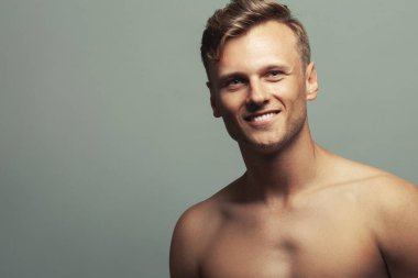 Erkek güzellik konsepti. Açık gri arka planda poz veren 30 yaşındaki gülümseyen sporcunun portresi. Kapatın. Kopyalama uzayı. Sporcu stili. Dalgalı, parlak sarı saçlar. Stüdyo çekimi