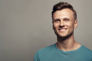 Erkek güzellik konsepti. Gri arka planda mavi tişörtlü gülümseyen 30 yaşındaki bir adamın portresi. Kapatın. Klasik bir tarz. Dalgalı, parlak sarı saçlar. Stüdyo çekimi