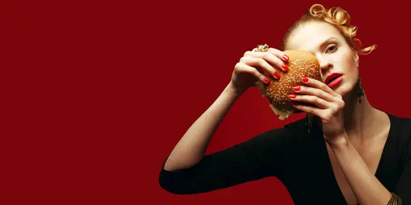 Fahion Fast Food Concept. Guilty pleasure. Fashionable model holding burger near face over red background. Perfect hair, skin, make-up, manicure. Golden accessories. Close up. Copy-space. Studio shot
