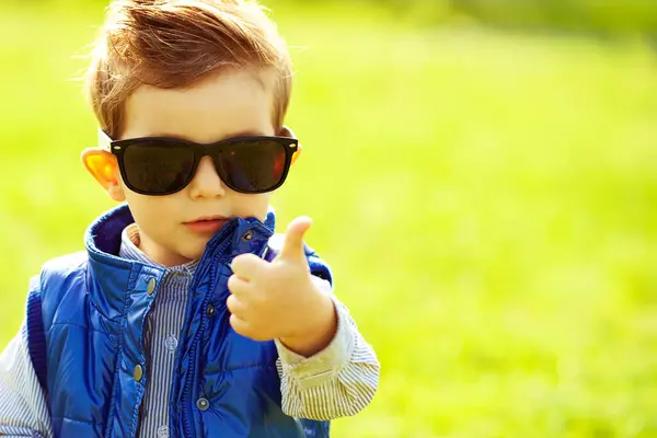 トレンディなサングラスと公園に立っている青いジャケットでショウガの髪のスタイリッシュな赤ちゃんの男の子は親指を示しています Okサイン ヒップスタースタイル 晴れた天気 コピースペース クローズアップ アウトドアショット ストック画像