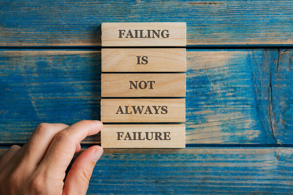 Male hand stacking five wooden pegs to spell the Failing is not a failure sign. Over textured blue wooden background.