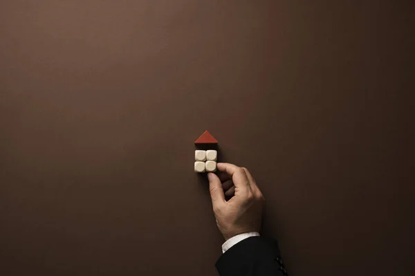 Hand of a businessman building a house of simple plain wooden blocks with a red roof. Over brown background with plenty of copy space.
