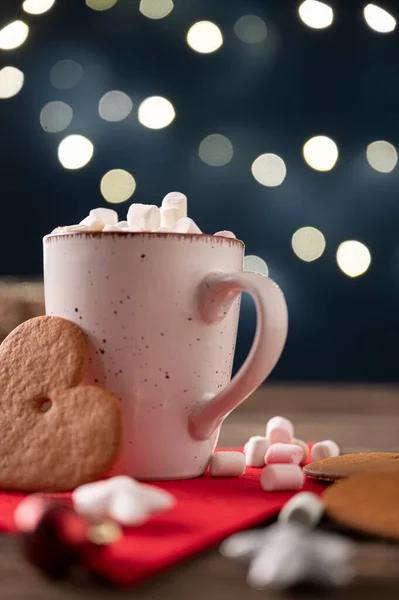 Cup of hot chocolate decorated with tiny marshmallows with a big cookie leaning on the mug. Over shiny background with christmas lights.