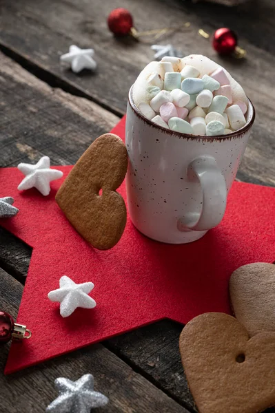 Cup of delicious hot chocolate with cream decorated with mini colorful marshmallows, placed on red star mat in a christmas setting and a big heart shaped cookie leaning on the mug.