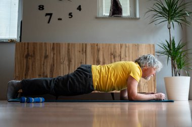 Senior man with grey curly hair in bright yellow tshirt exercising at home making a plank position. Conceptual image of senior life and ageing well.