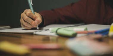 Low angle closeup view of a child making homework, writing in his notebook. Marker pens an pencils lying on a desk.