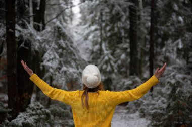 View from behind of a young woman in bright yellow sweater standing in the middle of peaceful snow covered winter forest with her arms spread widely.