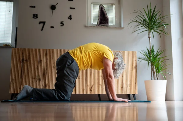 Male pensioner in yellow shirt making a pilates cat pose while working out and stretching on a mat at home. Active senior lifestyle.