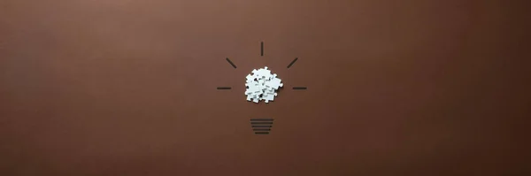 Wide view image of a light bulb made of scattered puzzle pieces and hand drawn shining rays. Over brown background.