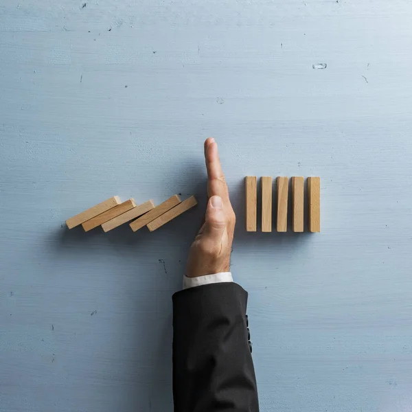 Overhead view of hand in a business suit stopping or intervening collapsing dominos. Over light blue background. Conceptual of crisis management and proactive solution.