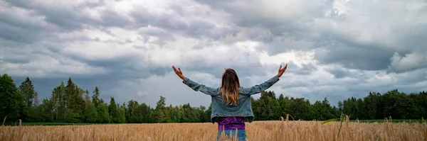 一位年轻女子站在金色的麦田里 张开双臂 向多云的夏日天空张开双臂 从身后看去 她在庆祝生命或沉思 — 图库照片