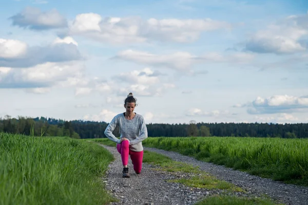 穿着粉红腿的真正的年轻女子伸展在美丽的绿色田野中间 在她去跑步前热身 — 图库照片