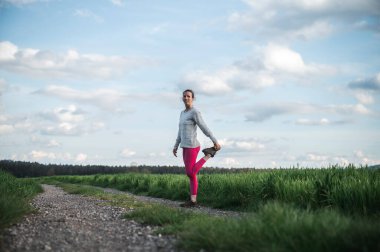 Pembe taytlı genç bir kadın koşuya çıkmadan önce ısınıyor ve ısınıyor. Güzel yeşil yay tarlalarının ortasında köy yolunda duruyor..