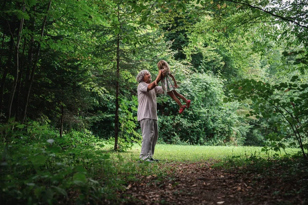 祖父と彼の孫娘の間の特別な瞬間 おじいちゃんは緑の夏の森の中で空気中で高いヒールの孫を持ち上げる — ストック写真