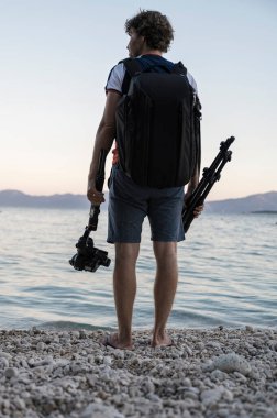 Genç, profesyonel bir fotoğrafçının, elinde tripodla, elinde kamerayla sakin bir denizin kıyısındaki güzel bir çakıl taşlı sahilde duruşu..