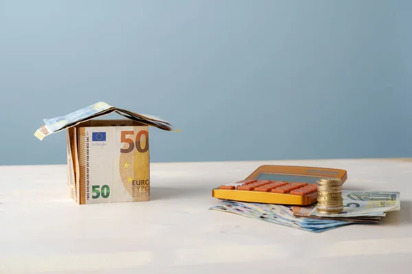 房子是由欧元钞票制成的 旁边是一个计算器和一堆钱 其概念上是房地产 抵押贷款和家庭保险 — 图库照片