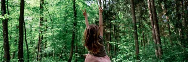 一个蹒跚学步的小女孩站在美丽的翠绿的森林里 双手高高地高高地举着胜利的姿态 从她身后看去 — 图库照片