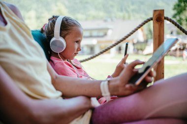 Modern teknoloji ve bağımlılık sorunları ve tuzakları - Kulaklıklı küçük bir kızın profil görüntüsü ve annesi bir kafede oturuyor ve birbirlerinden habersiz cep telefonları kullanıyor..