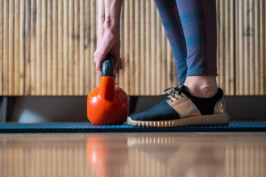 Kadın ellerinin portakal rengi bir kettlebell ağırlığı taşıdığı profil görüntüsü atletlerin ayaklarının evdeki güç egzersizlerinin kavramsal bir görüntüsüne yerleştirilmiş..