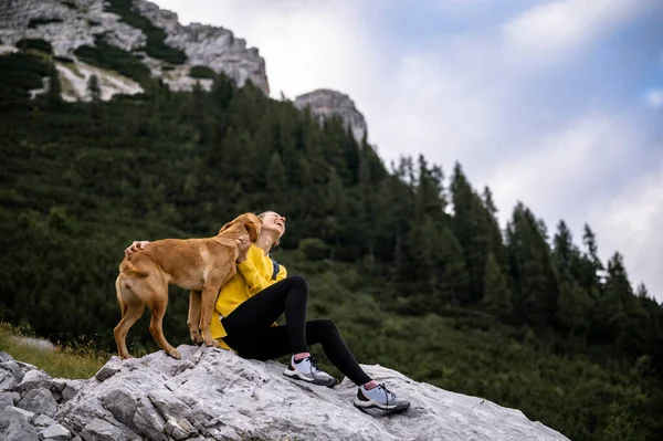 Pure and trustful relationship between a dog and a woman - golden labrador retriever dog kissing her owner sitting on a rock resting during a hike together.