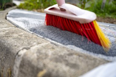 Yeni asfalt kaldırımın boşluklarına seramik kumu iten parlak kırmızı süpürgenin alçak açılı yakın çekim görüntüsü.