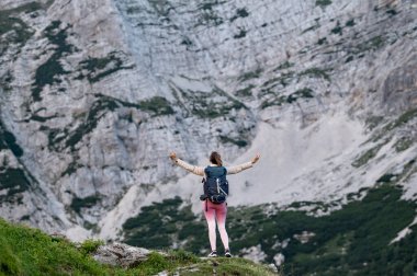 Sırt çantasıyla bir dağın tepesinde dikilen genç bir kadın yürüyüşçünün arkasından kutlamada diğer yüksek kayalık dağların manzarasına bakarken kollarını ardına kadar açar..
