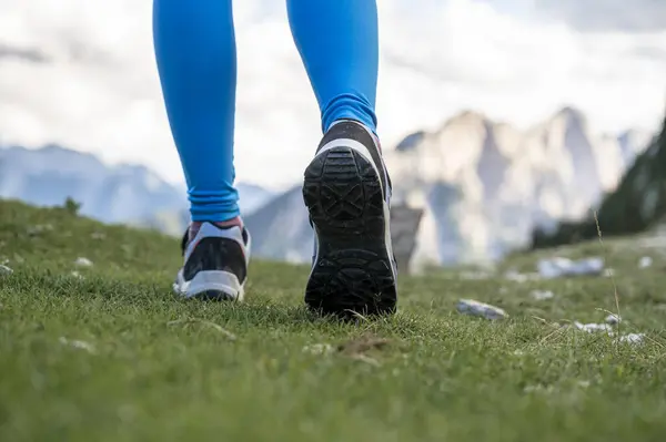 Low angle view of female legs in hiking shoes walking on green grass with high mountains in background. View of the sole of the shoes.