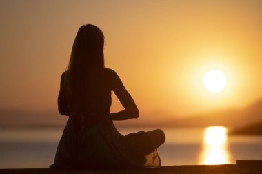 Yazlık elbiseli genç bir kadının, deniz kenarında, güzel, sıcak renkli gündoğumunda meditasyon yaparken yandan ve hafifçe arkasını görmek. Nilüfer pozisyonunda oturuyor, avuçlar göğsünün önünde birleşmiş..