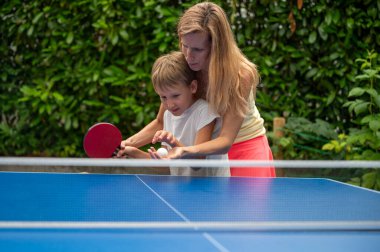 Genç anne küçük oğluna masa tenisi oynamayı ve raketi doğru tutmayı öğretiyor. Dışarıda doğal yeşil bir arka plan var., 