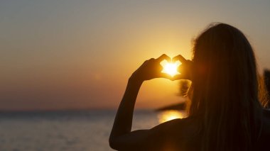 Omuz hizasında, parmaklarıyla kalp şekli oluşturan genç bir kadın vardı. Kalbinde parlayan sıcak turuncu güneş vardı. Deniz kenarında tatil, neşe ve özgürlüğün kavramsal bir görüntüsüyle. 