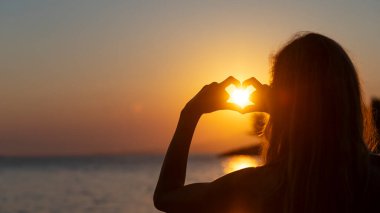 Omuz hizasında, parmaklarıyla kalp şekli oluşturan genç bir kadın vardı. Kalbinde parlayan sıcak turuncu güneş vardı. Deniz kenarında tatil, neşe ve özgürlüğün kavramsal bir görüntüsüyle. 