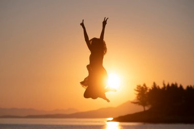 Deniz kenarındaki güzel gündoğumunda havaya zıplayan hayat dolu genç bir kadın. Yaz tatili viber kavramsal görüntü.