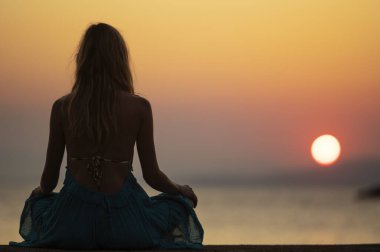 View from behind of a young womani n light summer dress sitting in lotus position by the sea at beautiful warm glowing sunrise. Summer vacation, peace and relaxation conceptual image.  clipart