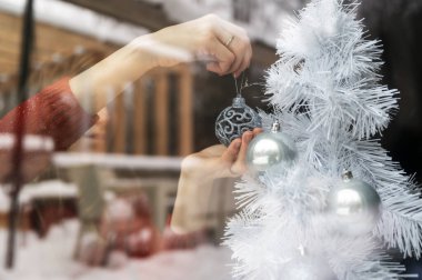 Beyaz Noel ağacını parlak mavi bir bayram mücevheriyle süsleyen genç bir kadının penceresinden bakın. Bardaktaki karlı doğanın yansımasıyla. 