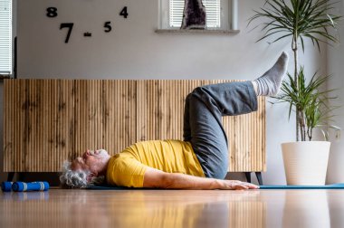 Sırtüstü uzanmış, bacakları havada, parlak sarı tişörtlü yaşlı bir adamın profil görüntüsü. Sağlıklı bir yaşam için güçlü çekirdek kasları için pilates yapıyorum.. 