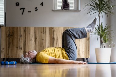 Sırtüstü uzanmış, bacakları havada, parlak sarı tişörtlü yaşlı bir adamın profil görüntüsü. Sağlıklı bir yaşam için güçlü çekirdek kasları için pilates yapıyorum.. 